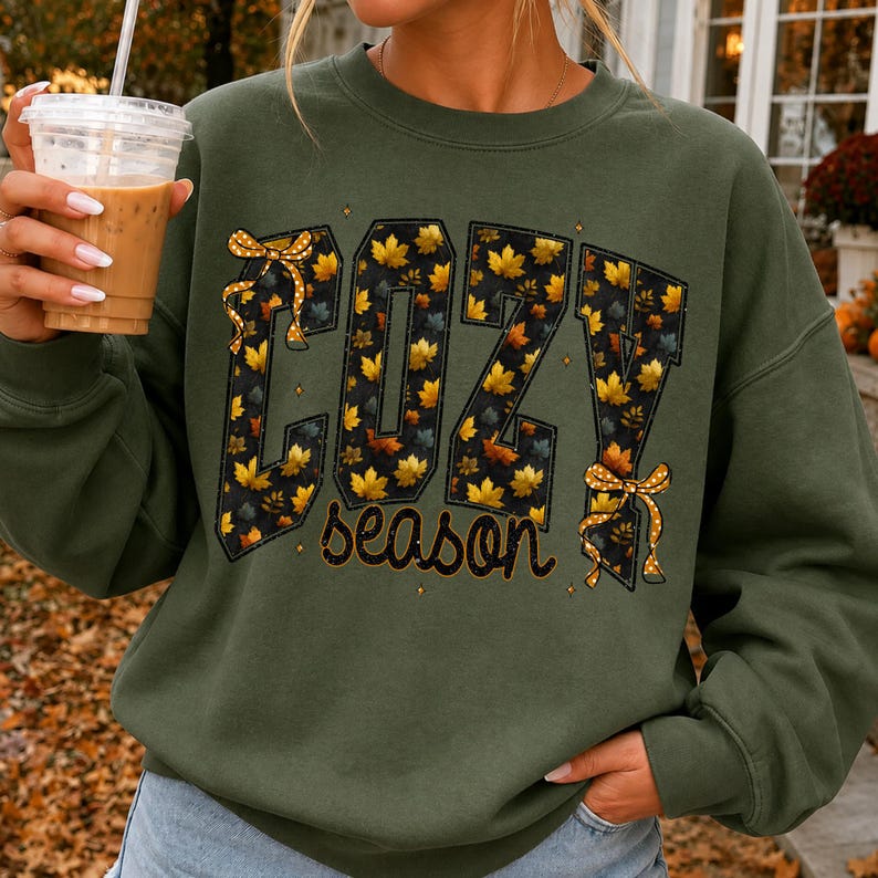 May include: An olive green crewneck sweatshirt featuring the text COZY season in a bold font. The letters are filled with a pattern of yellow and orange autumn leaves on a black background, accented by two yellow polka dot bows on the letters C and Y.