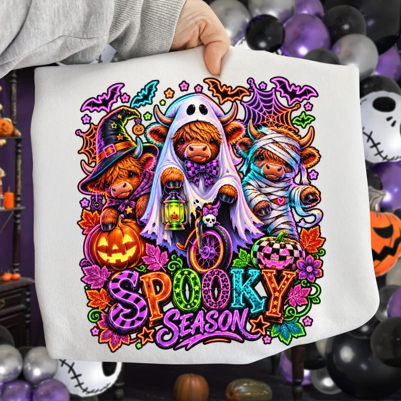 Może przedstawiać: Biała bluza z kolorową grafiką przedstawiającą trzy krowy rasy Highland przebrane na Halloween. Jedna ma kapelusz czarownicy, druga jest duchem z latarnią, a trzecia mumią. Napis Spooky Season wykonany jest żywymi, wzorzystymi literami.