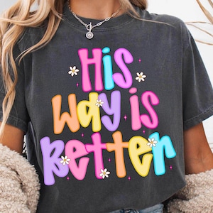 Può includere: T-shirt grigio scuro con la scritta "His Way Is Better" in lettere colorate. Il testo è impreziosito da piccoli fiori bianchi e stelle. La maglietta ha una vestibilità morbida.