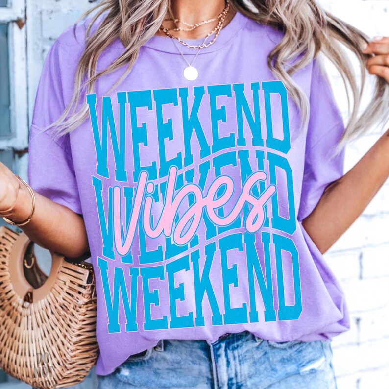 K&ouml;nnte beinhalten: Helllila T-Shirt mit dem Text "WEEKEND VIBES WEEKEND" in einem wellenf&ouml;rmigen Design. Der Text ist in Blau und Rosa gehalten. Das Shirt hat kurze &Auml;rmel und einen Rundhalsausschnitt.