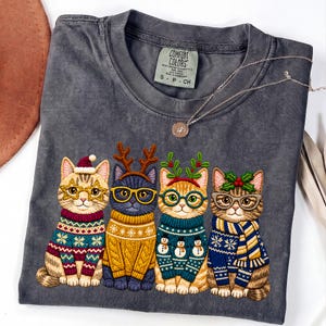 May include: A charcoal gray t-shirt featuring a graphic of four cats wearing holiday sweaters and glasses. The first cat wears a Santa hat, the second has reindeer antlers, the third has mistletoe, and the fourth has holly. A thin silver necklace rests on the collar.