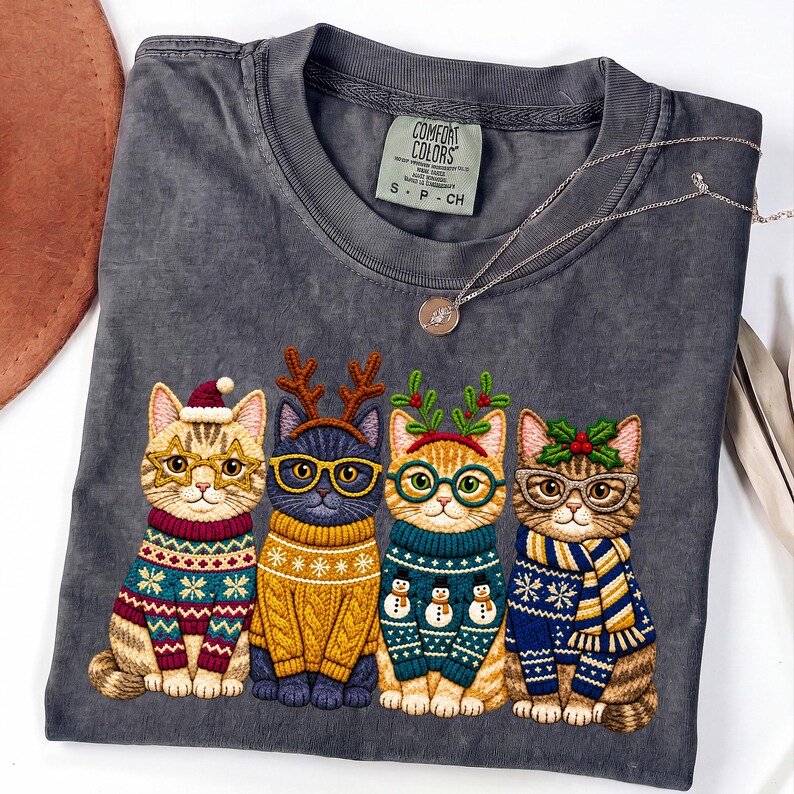 May include: A charcoal gray t-shirt featuring a graphic of four cats wearing holiday sweaters and glasses. The first cat wears a Santa hat, the second has reindeer antlers, the third has mistletoe, and the fourth has holly. A thin silver necklace rests on the collar.