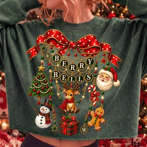Op de afbeelding: Een donkergroen sweatshirt met een grote afbeelding van een rode strik met stippen en hangende gouden kettingen. De tekst BERRY BELLS staat in cirkelvormige kaders. Versieringen omvatten een kerstboom, een sneeuwpop, een rendier en een cadeau.