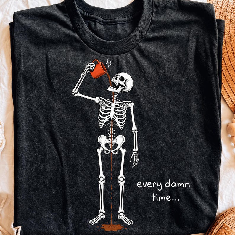 May include: A black crew neck t-shirt featuring a white skeleton graphic drinking from an orange mug. The coffee flows through the skeleton's ribcage and spills onto the floor. The text "every damn time..." is printed in white on the lower right side.