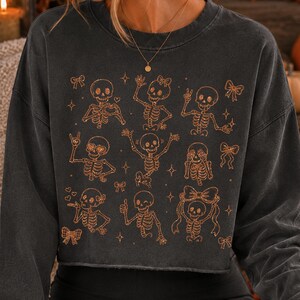 Op de afbeelding: Een antracietgrijze cropped sweater met een koperkleurige grafische print van meerdere skeletten in verschillende houdingen, zoals zwaaien, hartjes maken en strikjes dragen, afgewisseld met kleine ster- en striksymbolen.