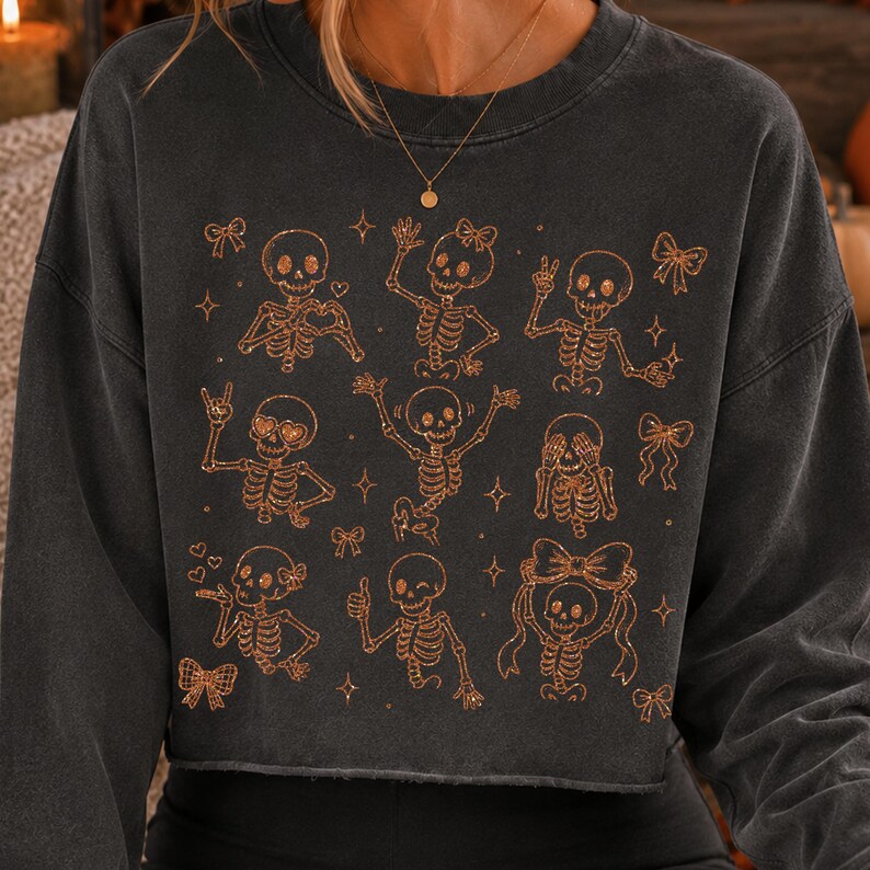 Op de afbeelding: Een antracietgrijze cropped sweater met een koperkleurige grafische print van meerdere skeletten in verschillende houdingen, zoals zwaaien, hartjes maken en strikjes dragen, afgewisseld met kleine ster- en striksymbolen.