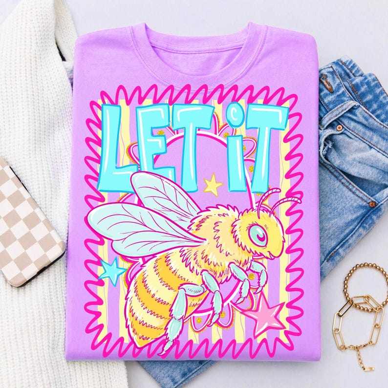 May include: A lilac-colored t-shirt featuring a cartoon bee with the words "LET IT" in turquoise. The bee is yellow and has light blue wings, surrounded by stars and a pink wavy border. The design is set against a striped background.