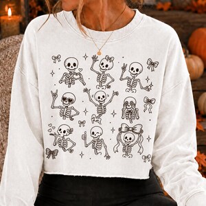 Op de afbeelding: Een wit, langmouwig, kort sweatshirt met een zwarte lijntekening-print van meerdere skeletten in verschillende houdingen, waaronder zwaaiend, knipogend en hartvormen makend, afgewisseld met kleine ster- en strikillustraties.