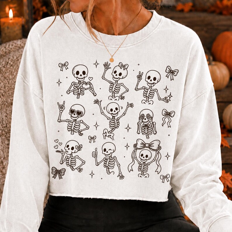 Op de afbeelding: Een wit, langmouwig, kort sweatshirt met een zwarte lijntekening-print van meerdere skeletten in verschillende houdingen, waaronder zwaaiend, knipogend en hartvormen makend, afgewisseld met kleine ster- en strikillustraties.