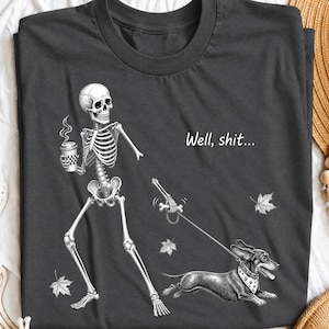 May include: A dark gray t-shirt featuring a white graphic of a skeleton holding a coffee cup and walking a dachshund on a leash. The text "Well, shit..." is printed above the dog, accompanied by small white illustrations of falling autumn leaves.