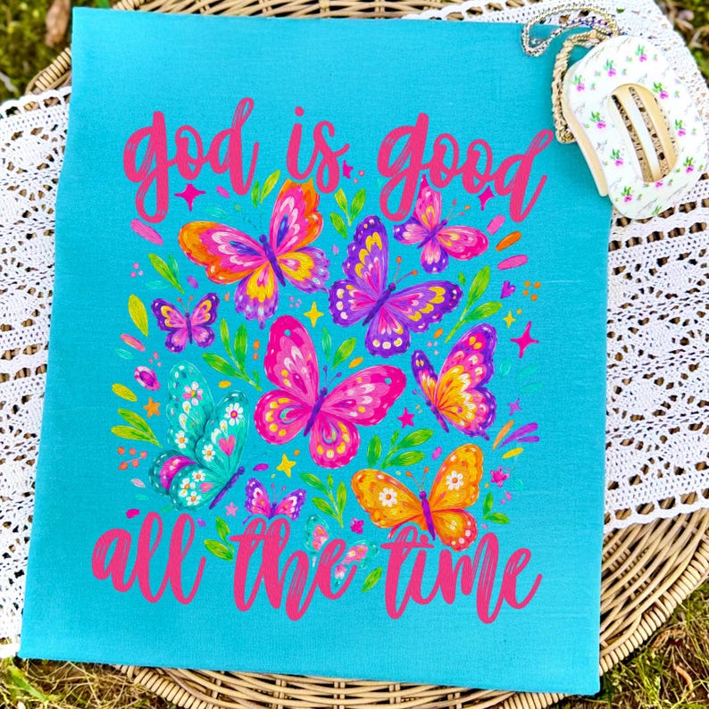 Peut inclure: Un sac fourre-tout en toile turquoise avec un motif de papillons vibrants et le texte rose "God is good all the time". Les papillons sont dans des tons de rose, orange, jaune et violet.