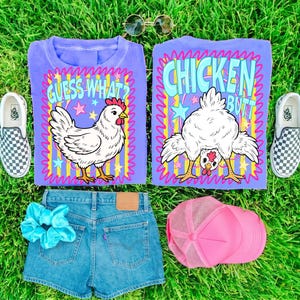 May include: A flat lay featuring a purple t-shirt with a cartoon chicken graphic and the text "Guess What?" and "Chicken Butt". Also included are denim shorts, a pink trucker hat, a blue scrunchie, and checkered slip-on shoes, all on a green grassy surface.