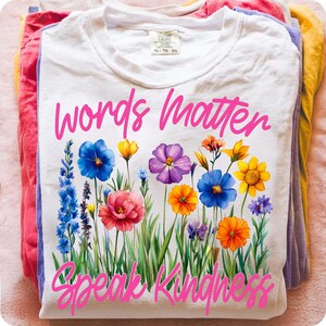 Puede incluir: Camiseta blanca con el texto "Words Matter Speak Kindness" en rosa. La camiseta presenta un dise&ntilde;o floral colorido con flores azules, amarillas, moradas y naranjas. La camiseta est&aacute; doblada encima de otras camisetas de colores.