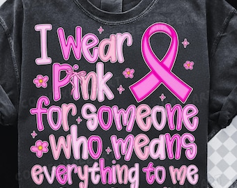 以下が含まれることがあります： 「I Wear Pink for someone who means everything to me」のピンク色の文字がプリントされた濃いグレーのTシャツ。ピンクのリボンと花のアクセントも特徴です。このシャツは乳がん啓発を促進しています。
