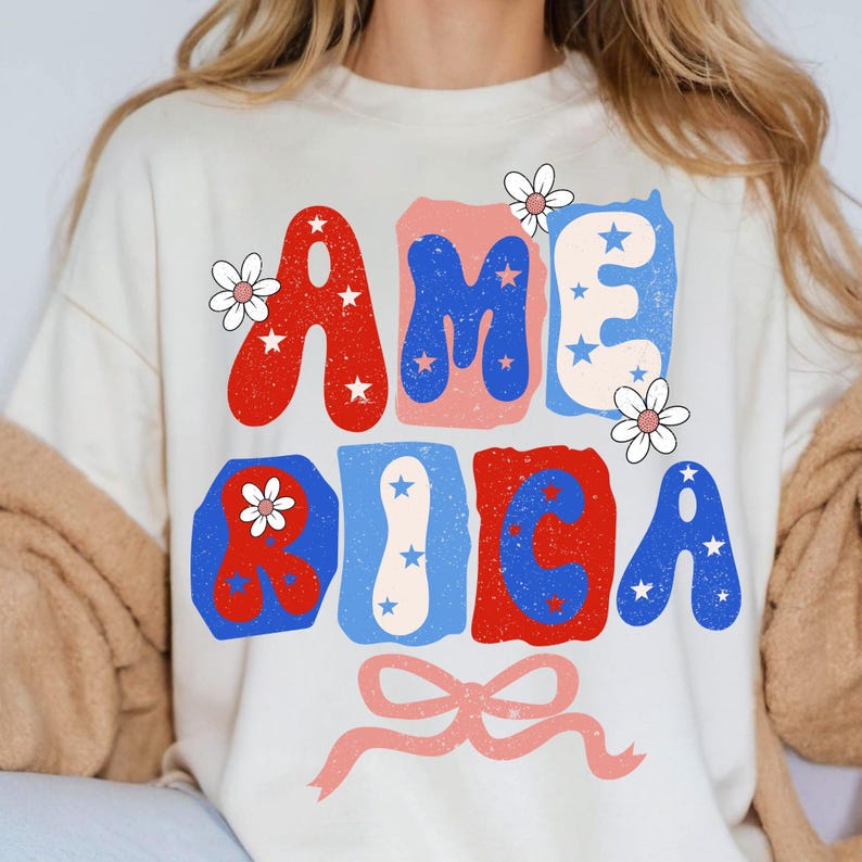 May include: A cream-coloured sweatshirt featuring the word "AMERICA" in red, blue, and light pink bubble letters. The letters are decorated with white stars and small white flowers. A light pink bow is at the bottom.