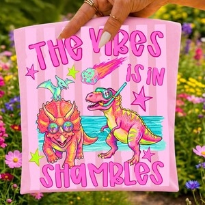 Peut inclure: T-shirt rose avec l'inscription "THE VIBES IS IN SHAMBLES" et un graphisme de dinosaures de dessins anim&eacute;s, d'une m&eacute;t&eacute;orite et d'&eacute;toiles. Les dinosaures portent des lunettes de soleil et un tuba. Le fond est &agrave; rayures roses.