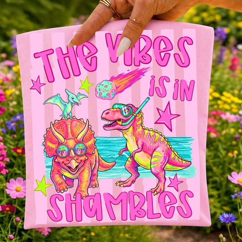 Peut inclure: T-shirt rose avec l'inscription "THE VIBES IS IN SHAMBLES" et un graphisme de dinosaures de dessins anim&eacute;s, d'une m&eacute;t&eacute;orite et d'&eacute;toiles. Les dinosaures portent des lunettes de soleil et un tuba. Le fond est &agrave; rayures roses.