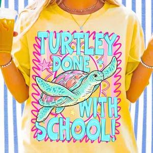 以下が含まれることがあります： カラフルなウミガメのグラフィックと「TURTLEY DONE WITH SCHOOL！」の文字が入った黄色のTシャツ。カメはターコイズブルーの体とピンクとオレンジの甲羅をしています。テキストは青とピンクです。