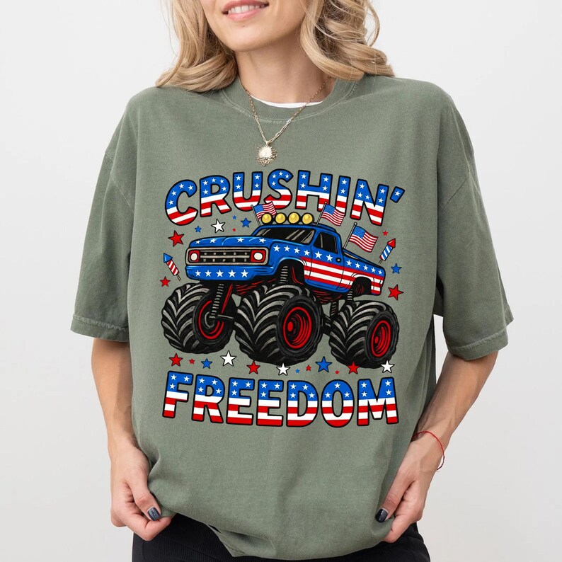 K&ouml;nnte beinhalten: Salbeigr&uuml;nes T-Shirt mit patriotischem Design. Die Grafik zeigt einen blauen Monster Truck mit roten und wei&szlig;en Akzenten, die Worte "CRUSHIN' FREEDOM" in Rot, Wei&szlig; und Blau sowie Sterne und Streifen.