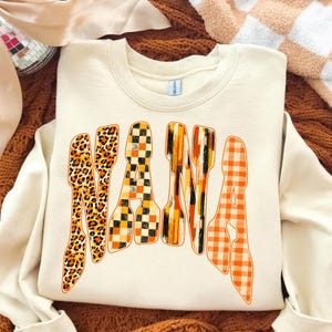 May include: A cream-colored crewneck sweatshirt featuring the word NANA printed in large, arched letters. Each letter displays a distinct pattern: leopard print, black and orange checkerboard, vertical orange and black brushstrokes, and orange gingham.