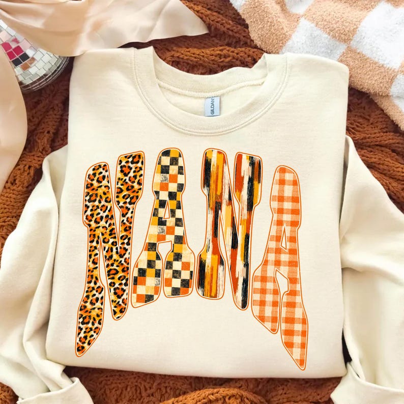 May include: A cream-colored crewneck sweatshirt featuring the word NANA printed in large, arched letters. Each letter displays a distinct pattern: leopard print, black and orange checkerboard, vertical orange and black brushstrokes, and orange gingham.