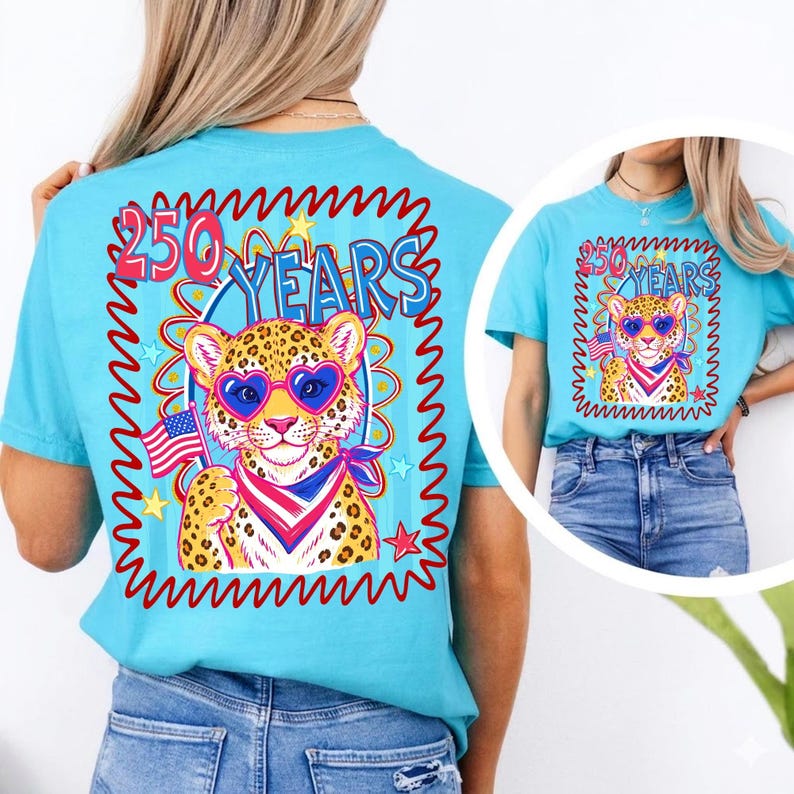 Op de afbeelding: Een lichtblauw t-shirt met een afbeelding van een luipaard met een hartvormige zonnebril en een patriottische bandana, met een Amerikaanse vlag. De tekst "250 YEARS" staat boven de luipaard in een golvend kader.