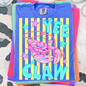 May include: A stack of colorful t-shirts. The top shirt is blue with a graphic of a lobster wearing a cowboy hat, the words "YEE CLAW", and star accents. The shirt has yellow vertical stripes. Other shirts are pink, purple, and orange.
