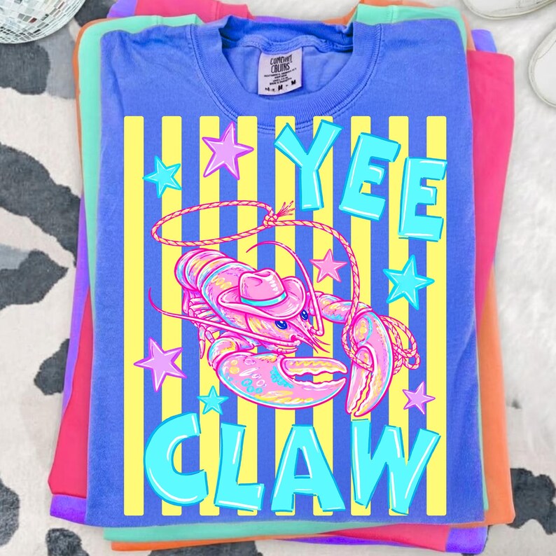 May include: A stack of colorful t-shirts. The top shirt is blue with a graphic of a lobster wearing a cowboy hat, the words "YEE CLAW", and star accents. The shirt has yellow vertical stripes. Other shirts are pink, purple, and orange.