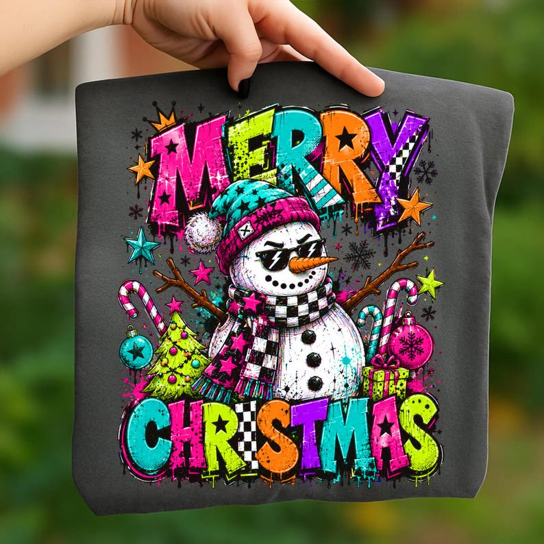 Puede incluir: Una camiseta gris oscuro con un gr&aacute;fico de un mu&ntilde;eco de nieve con gafas de sol, un gorro y una bufanda a cuadros. El texto MERRY CHRISTMAS est&aacute; impreso en letras grandes y coloridas estilo grafiti. El dise&ntilde;o incluye bastones de caramelo.