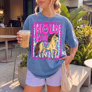 May include: A blue t-shirt with the words "HOLD YOUR" in pink block letters and a cartoon horse graphic. The horse has a saddle and is surrounded by stars and a pink zig-zag border. The shirt is worn with striped shorts.