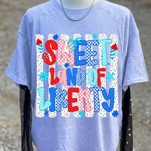 May include: A light gray t-shirt featuring a graphic print with the text SWEET LAND OF LIBERTY in red, white, and blue checkered and polka dot patterns. The design includes firework illustrations and small blue flowers on a vertical striped background.