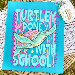 以下が含まれることがあります： ターコイズ色のTシャツ。カラフルなカメのグラフィックと「TURTLEY DONE WITH SCHOOL！」の文字。カメはピンクとオレンジ色の甲羅を持ち、ピンクと青のアクセントで囲まれています。楽しくお祝いのデザインです。