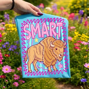 May include: A light blue t-shirt featuring a cartoon bison with the word "SMART" in pink bubble letters. The bison is tan with a shaggy mane and horns. The design includes stars and wavy lines.
