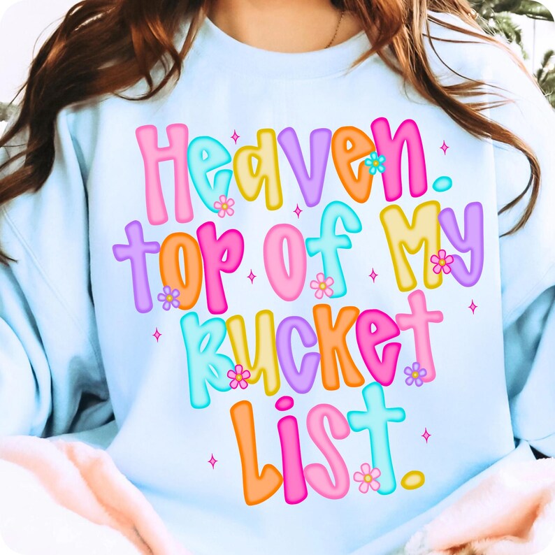 K&ouml;nnte beinhalten: Hellblaues Sweatshirt mit dem Text "Heaven, Top of My Bucket List" in bunten Buchstaben. Der Text ist mit kleinen Blumen und Sternen verziert.
