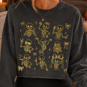 Op de afbeelding: Een antracietgrijze cropped sweater met een gouden glitter grafische print van negen verschillende skeletten in diverse houdingen, zoals zwaaien, hartjes maken en strikjes dragen, afgewisseld met kleine gouden sterretjes en strikjes.