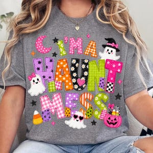 May include: A heather gray t-shirt featuring the text "C'I'M A HAUNT MESS" in colorful, patterned block letters. The design includes three white ghosts, a pink jack-o'-lantern bucket filled with candy corn, and small black stars.