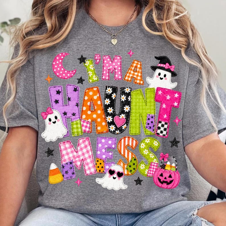 May include: A heather gray t-shirt featuring the text "C'I'M A HAUNT MESS" in colorful, patterned block letters. The design includes three white ghosts, a pink jack-o'-lantern bucket filled with candy corn, and small black stars.
