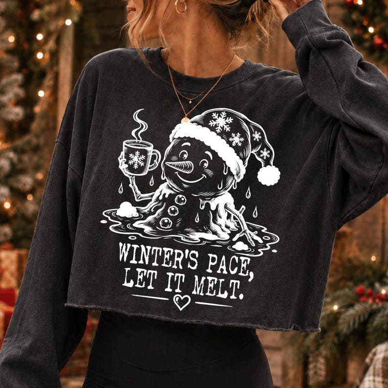 May include: A charcoal gray long-sleeve cropped sweatshirt featuring a white graphic of a melting snowman wearing a Santa hat and holding a steaming mug. The text reads, "WINTER'S PACE, LET IT MELT." with a small heart icon below the lettering.