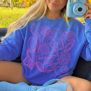 K&ouml;nnte beinhalten: Ein blaues Sweatshirt mit einem rosa Muscheldesign. Die Grafik zeigt verschiedene Muschelillustrationen, wie z. B. Muscheln, Jakobsmuscheln und Seesterne. Das Sweatshirt hat eine einfarbige blaue Farbe.