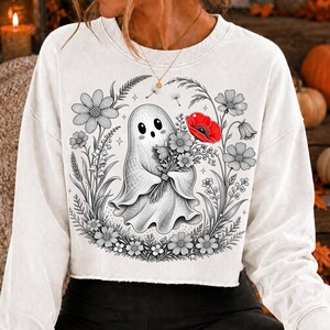 K&ouml;nnte beinhalten: Ein wei&szlig;es Langarm-Sweatshirt mit einer zentralen Grafik eines Geistes, der einen Blumenstrau&szlig; h&auml;lt. Die Illustration ist haupts&auml;chlich in Schwarz-Wei&szlig; gehalten, mit einer einzelnen leuchtend roten Mohnblume. Der Geist ist von Blumen umgeben.