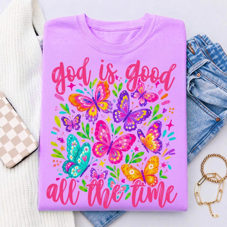 Peut inclure: T-shirt lilas avec un motif de papillons color&eacute;s et l'inscription rose "God is good all the time". Les papillons sont dans les tons de rose, orange, violet et turquoise.