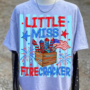 May include: A light gray t-shirt with the words "LITTLE MISS FIRECRACKER" in red and blue. The shirt features a graphic of fireworks, an American flag, and stars, perfect for Independence Day celebrations.