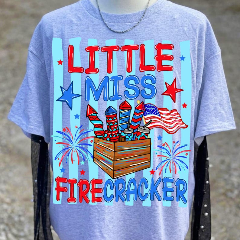 May include: A light gray t-shirt with the words "LITTLE MISS FIRECRACKER" in red and blue. The shirt features a graphic of fireworks, an American flag, and stars, perfect for Independence Day celebrations.