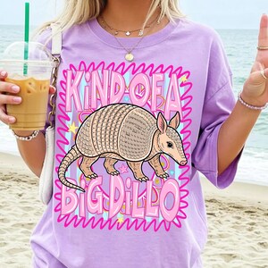 May include: A lilac t-shirt featuring the words "KIND OF A BIG DILLO" and an armadillo illustration. The armadillo is depicted in tan and pink, with the text in pink and blue. The shirt has short sleeves.