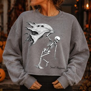 K&ouml;nnte beinhalten: Ein graumeliertes Sweatshirt mit Rundhalsausschnitt und einer schwarz-wei&szlig;en Grafik eines Skeletts, das ein Geisterlaken h&auml;lt. Das Laken ist mit einem Himmelsmuster aus Sternen und Monden versehen. Der Text "Sheet Happens.." steht unter dem Skelett.