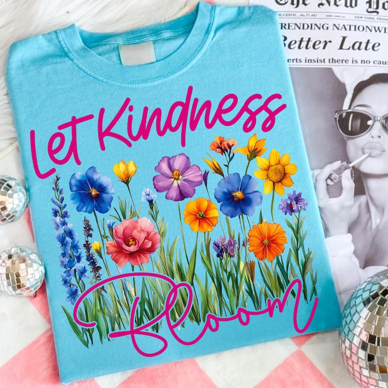 May include: A light blue t-shirt with the words "Let Kindness Bloom" in pink script. The shirt features a colorful floral design with various flowers in shades of blue, pink, yellow, and orange.