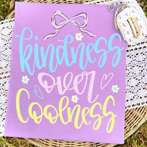 May include: A lavender t-shirt with the words "Kindness Over Coolness" in blue, pink, and yellow script. The design includes a bow, hearts, and flower accents. A small floral purse with a gold chain is in the upper right corner.