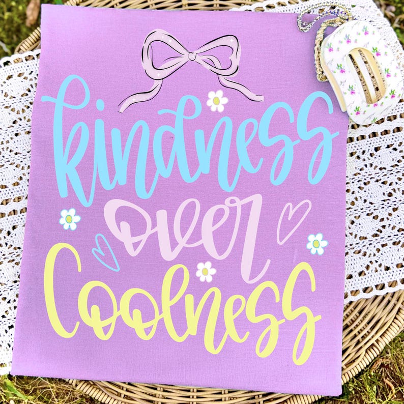 May include: A lavender t-shirt with the words "Kindness Over Coolness" in blue, pink, and yellow script. The design includes a bow, hearts, and flower accents. A small floral purse with a gold chain is in the upper right corner.