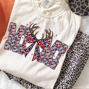 Peut inclure: T-shirt cr&egrave;me avec le mot "MAMA" dans un motif camouflage rouge, blanc et bleu. Un n&oelig;ud en bois de cerf marron est centr&eacute; au-dessus du mot. Un pantalon de pyjama &agrave; imprim&eacute; l&eacute;opard est visible sur le c&ocirc;t&eacute;.