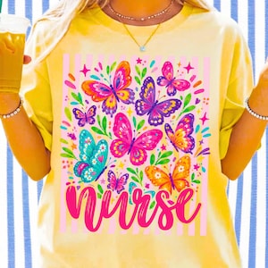 May include: A pale yellow t-shirt featuring a central graphic of colorful butterflies in shades of pink, orange, purple, and teal, accented with green leaves and small stars. The word nurse is printed in a bold, cursive pink font at the bottom.